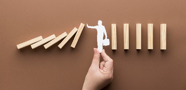 a hand holding a cutout of a corporate person standing between dominoes and stopping them from falling onto other dominoes